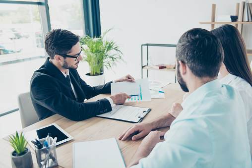 Rear view of couple sitting in the brunet`s smart lawyer`s work station,  discussing the possibility f taking credit for buying purchase, all are dressed in formal outfits